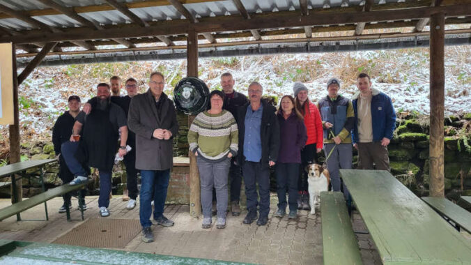 Landrat Dietmar Seefeldt (Fünfter von links) mit Engagierten des Wanderclubs Nello e.V. am AED-Gerät, das unter einem Unterstand an der Nello-Hütte befestigt und dadurch leicht zugänglich ist. (Foto: KV SÜW)